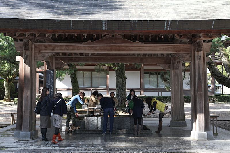 Visitante de un templo japonés realizando un ritual tradicional de purificación en la fuente Temizuya, lavándose las manos con un cucharón de madera en preparación para la oración y la visita al templo.