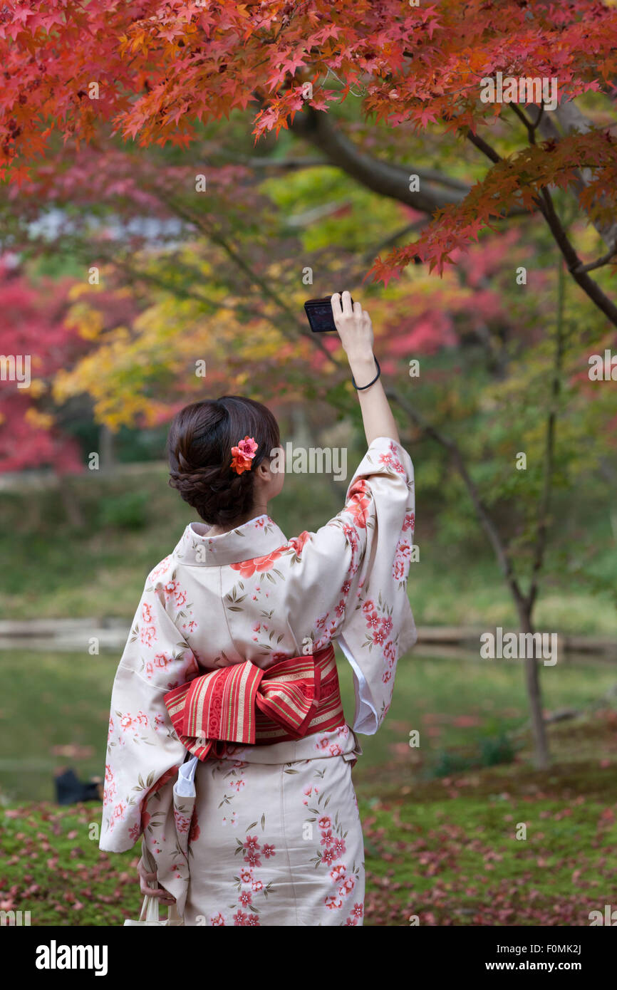 Una joven japonesa con un kimono tradicional fotografía brillantes hojas de arce otoñales, mostrando las oportunidades de fotografía del follaje otoñal de Kioto.